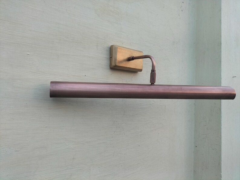 May include: A wall-mounted picture light with a long, cylindrical, metallic copper-toned shade. The fixture features a rectangular brass-colored base plate and a thin, curved arm that connects the base to the center of the horizontal light tube.