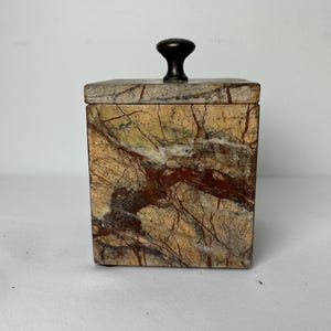 May include: A square stone canister featuring a polished finish with natural veining in shades of tan, brown, and cream. The container includes a matching square lid topped with a small, dark brown, rounded knob handle centered on top.