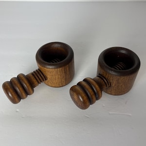 May include: Two dark brown wooden nutcrackers featuring a cylindrical body with a hollow center and a threaded screw mechanism. Each tool has a rounded handle with three distinct horizontal ridges, showcasing a smooth, polished wood grain finish.