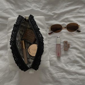 May include: A white corduroy cosmetic bag with black lace trim, open to show makeup items. Gold-framed sunglasses, a lip gloss, and a hair clip are arranged on a white surface.