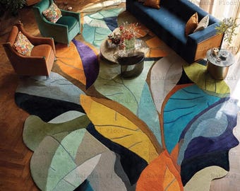 May include: A large, leaf-shaped area rug with a colorful design. The rug features shades of orange, yellow, green, blue, and purple. The rug is placed on a hardwood floor, and the room includes furniture like a blue sofa and armchairs.