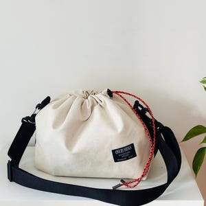 May include: A beige drawstring bag with a black adjustable shoulder strap. The bag has a red and white rope drawstring closure and a small black label that reads "ONE BY SHOKS".