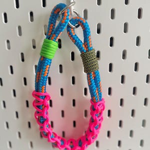 May include: A vibrant dog collar made from blue and orange rope, with green and black detailing. The collar has a bright pink decorative section. A silver metal ring is at the top.