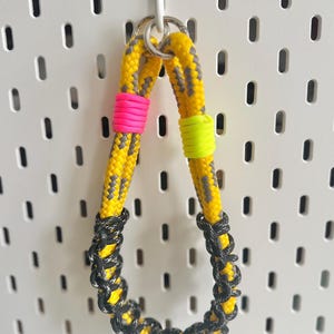 May include: A yellow and grey rope keychain with a silver ring. The keychain features pink and neon green accents and a black and grey woven loop. It hangs against a white pegboard.