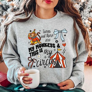 A woman wearing a gray sweatshirt with a graphic design of a monkey and circus-related elements, holding a white mug.