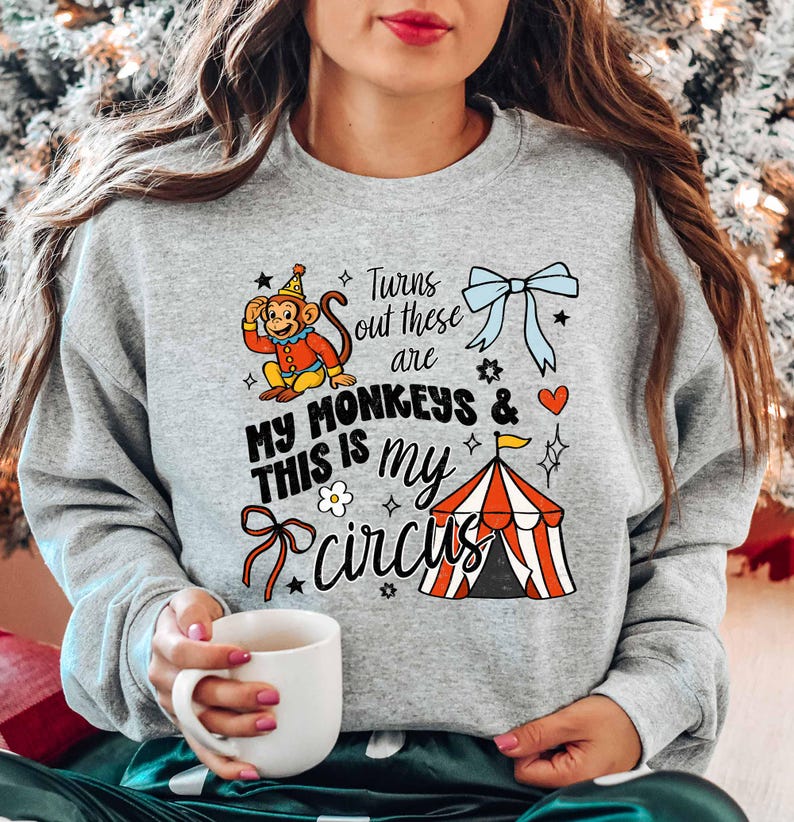 A woman wearing a gray sweatshirt with a graphic design of a monkey and circus-related elements, holding a white mug.