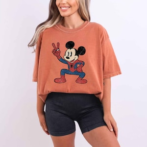 A woman wearing a red t-shirt with a cartoon character and black shorts is smiling.