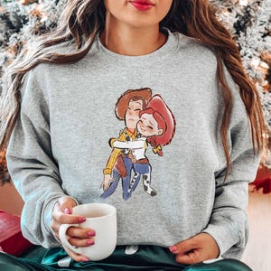A woman wearing a gray sweatshirt with a cartoon character printed on it, holding a white mug in her hand.