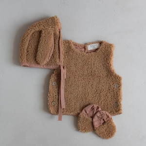 Op de afbeelding: Een driedelige baby set in een teddybeer thema. De set bevat een bruin vest met knoopsluitingen, een bijpassende muts met oordetails en een paar babyslofjes. De set is gemaakt van een zacht, pluche materiaal.