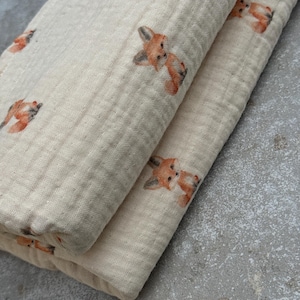 Op de afbeelding: Gevouwen beige mousseline stof met een herhaald patroon van oranje en bruine vos illustraties. De stof heeft een getextureerde, gekreukelde uitstraling. De vos illustraties zijn gedetailleerd met zichtbare ogen, oren en vacht.