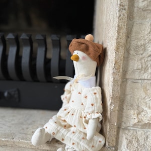 Puede incluir: Muñeca de tela hecha a mano, cuerpo blanco, con boina marrón y vestido crema con detalles florales marrones. La muñeca tiene pico amarillo y está sentada en una repisa de piedra cerca de una chimenea.