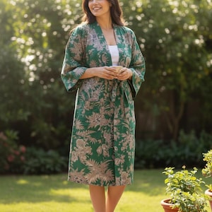 May include: A green floral print robe with a matching tie, worn over a white top. The robe features a botanical design in shades of green and tan. The model is standing barefoot in a garden setting, with potted plants in the foreground.