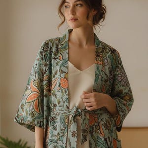 Puede incluir: Una bata tipo kimono con estampado floral azul claro con cinturón a juego. La bata presenta un vibrante estampado de flores y hojas naranjas, verdes y moradas. La modelo lleva una camiseta sin mangas y pantalones color crema. La bata tiene mangas anchas.