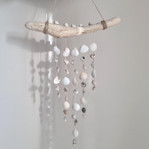 May include: A decorative wall hanging made from driftwood and seashells. The driftwood is a light brown colour, with small shells attached. Strands of white and beige seashells hang below, creating a mobile. The shells vary in size and shape.