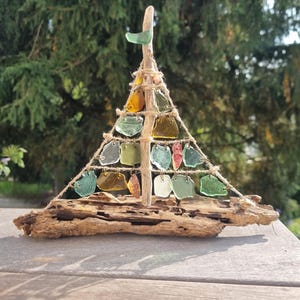 Könnte beinhalten: Eine dreieckige Skulptur aus einem Treibholzsockel und einem vertikalen Treibholzstamm. Schnüre bilden eine Baumform, die mit verschiedenen kleinen, glatten, durchscheinenden Meerglasstücken in Grün, Bernstein und klaren Farben verziert ist.
