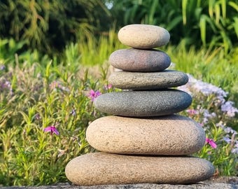 May include: A stack of six smooth, oval-shaped stones, balanced atop one another. The stones vary in color from light tan to gray and purple. The background features green foliage and small purple flowers, suggesting a natural outdoor setting.