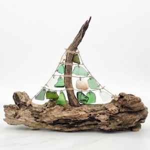 May include: A decorative sailboat sculpture crafted from a large piece of driftwood as the base and mast. The sail is constructed from a triangular arrangement of twine featuring various shades of green sea glass and two small white seashells.