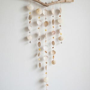 Può includere: Una decorazione da parete con un ramo di legno naturale, conchiglie e perline. Le conchiglie sono di varie forme e dimensioni, con tonalità neutre. Le perline sono trasparenti, gialle e grigie. Il pezzo è sospeso da spago naturale.