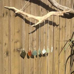 Könnte beinhalten: Treibholz- und Meeresglas-Mobile. Ein natürlicher Ast aus Holz hält Stränge aus Schnur, an denen jeweils ein Stück farbiges Meeresglas hängt. Die Meeresglasstücke haben verschiedene Formen und Farben, darunter Grün, Orange und Klar.