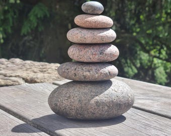 May include: A vertical stack of six smooth, rounded granite stones arranged in descending order of size. The stones feature a speckled texture with natural shades of gray, tan, and pinkish-brown, balanced carefully to form a single column.