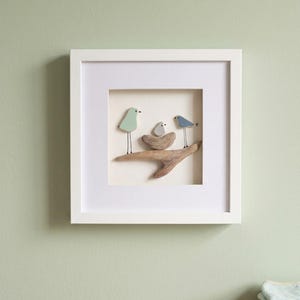 Könnte beinhalten: Ein quadratisches, weiß gerahmtes Wandbild mit einer Kieselstein-Szene. Drei Vögel sind aus glatten Steinen und dünnen Drahtbeinen gefertigt und sitzen auf einem Stück Treibholz. Ein Vogel ist hellgrün, einer blau und ein kleiner grauer sitzt im Nest.