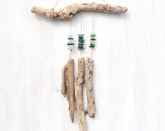 May include: A wall hanging featuring a horizontal piece of driftwood suspended by a thin cord. Three vertical strands hang from the driftwood, each decorated with small, tumbled green and clear glass pieces above a larger piece of driftwood.