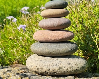 May include: A vertical stack of six smooth, rounded river stones of varying sizes and colors, including shades of gray, tan, and reddish-brown, balanced on top of a larger, textured, lichen-covered rock in an outdoor setting.