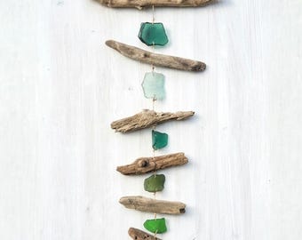 May include: A vertical wall hanging featuring five pieces of smooth sea glass in various shades of green and five pieces of weathered driftwood. The components are connected by thin twine, suspended from a larger horizontal piece of driftwood.
