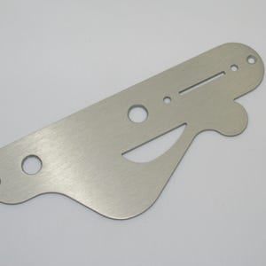 May include: A flat, brushed silver-coloured metal control plate for a musical instrument. The plate features an elongated, irregular shape with rounded edges, two circular holes, one small circular hole, and a long, narrow rectangular slot.