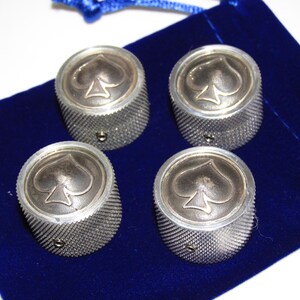 May include: A set of four cylindrical metal knobs with a textured, knurled side surface and a smooth top featuring an embossed spade symbol. Each knob includes a small side screw for attachment, presented on a deep blue velvet fabric pouch.