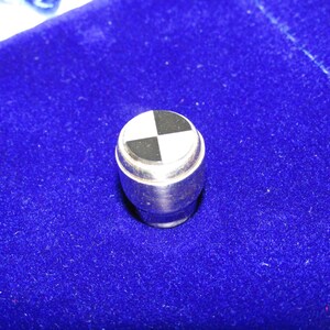 May include: A cylindrical metal knob featuring a circular top inlay with a black and white checkered pattern. The metallic body has a smooth, polished finish, and the top surface is divided into four equal quadrants of alternating black and white colors.