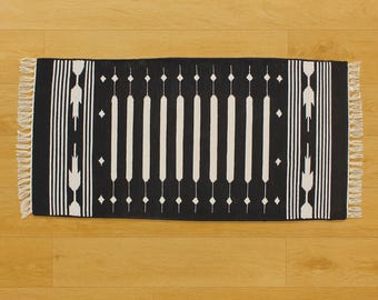 May include: A rectangular black and white patterned rug with a geometric design. The rug features vertical white stripes and diamond shapes on a black background. It has fringed edges and is displayed on a wooden floor.