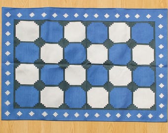 May include: A rectangular blue and white patterned rug. The rug features a checkerboard design with alternating blue and white squares. A border of blue with white diamond-shaped accents surrounds the central pattern.