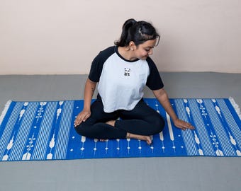 May include: A blue yoga mat with white vertical stripes and decorative floral patterns. The mat has a fringed edge. The person is wearing a black and white shirt with a panda graphic and black pants.