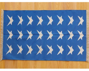 May include: A rectangular blue rug with a repeating pattern of white star shapes. The rug has a fringed edge on the short sides. The rug is displayed on a wooden floor.