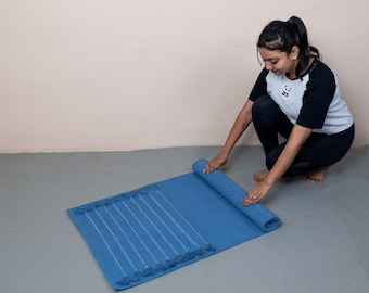 Op de afbeelding: Een blauwe yogamat met witte strepen en franjes wordt opgerold. De mat ligt op een grijze vloer, met een lichtbeige muur op de achtergrond. De mat lijkt gemaakt van een zacht, geweven materiaal en is ongeveer 173 cm lang.