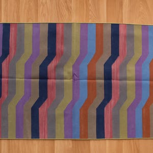 May include: Rectangular rug with a geometric pattern of vertical stripes in shades of green, purple, blue, and orange. The rug has fringed edges and is displayed on a wooden floor. A decorative floor covering.