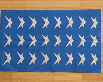 May include: A blue rectangular rug with a repeating pattern of white star shapes. The rug has a fringed edge on the top and bottom. The rug is lying on a wooden floor.