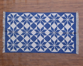May include: A rectangular blue and white patterned rug with a geometric starburst design. The rug has a blue border and fringe on the short ends. The rug is placed on a wooden floor.