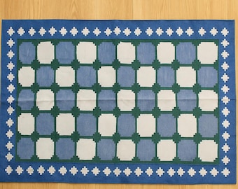 May include: A rectangular rug with a blue border and a geometric pattern. The pattern features alternating squares of white, light blue, and green. The border is adorned with white cross-shaped designs.