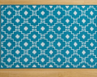May include: A rectangular turquoise rug with a white geometric pattern. The design features repeating diamond shapes and angular lines. The rug is placed on a light brown wooden floor.