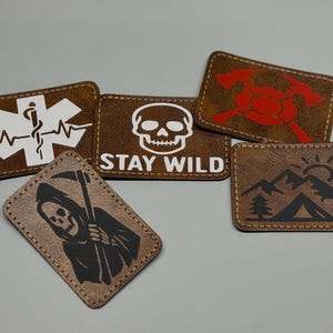May include: A collection of brown rectangular patches with various designs. One patch features a white medical symbol, another has a skull with "STAY WILD" text, and others display a Grim Reaper, axes, and a mountain scene.