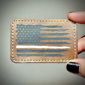 May include: A rectangular patch with a distressed American flag design. The flag features a brown background, blue stripes and stars, and a white stripe. The patch is held by a hand and has a stitched border.