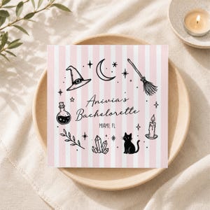 May include: A square paper napkin with a light pink and white vertical striped pattern. The center features black line art including a witch hat, crescent moon, broom, potion bottle, crystals, a cat, a candle, and text reading Anivia's Bachelorette MIAMI, FL.