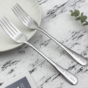 May include: Two stainless steel forks with the text "CUSTOM TEXT HERE" engraved on the tines and handles. The forks are placed on a white plate and a light-colored wooden surface. A sprig of greenery is in the background.
