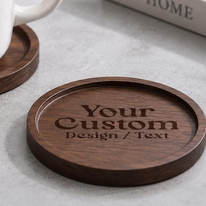 Puede incluir: Un posavasos circular de madera de color marrón oscuro con un borde elevado y un acabado liso. El centro del posavasos presenta el texto grabado "Your Custom Design / Text" en una fuente serif. La veta de la madera es visible en la superficie.
