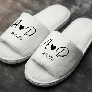 Peut inclure: Deux pantoufles blanches en tissu éponge. Chaque pantoufle porte les initiales "A" et "D", un symbole de cœur et la date "10.10.2026" en noir. Les pantoufles ont un design ouvert et une texture douce.