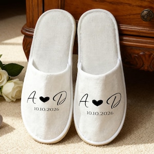 Op de afbeelding: Een paar witte, zachte slippers met een gepersonaliseerd ontwerp. Elke slipper heeft de initialen "A" en "D" met een hartsymbool en de datum "10.10.2026" in zwarte letters. De slippers staan op een neutrale achtergrond.