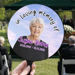 Pode incluir: Um leque comemorativo redondo com o retrato de uma pessoa de cabelo branco curto e blusa preta. O texto "in loving memory of" está impresso a preto, com o nome "Susan Baker" e as datas "03.06.1958 - 06.21.2026" em roxo.
