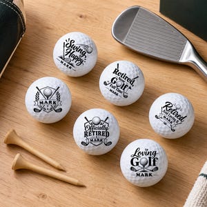 May include: Six white golf balls with black printed text and graphics, including the name Mark, arranged with two wooden tees, a metal golf club, a dark green box with text, and a black leather golf bag accessory on a light wood surface.